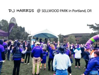 A picture of DJ Aaron Harris performing at the End Lupus Now event at Sellwood Park in Portland Oregon. The picture is taken from the back of the crowd facing the performance stage.