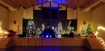 A picture of our DJ setup for a high school winter formal dance at Philomath Scout Lodge in Philomath, OR. There are multiple holiday trees of varying sizes strung with white lights on the stage. There are 4 blue colored led dance floor lights positioned in front of the DJ setup to add a splash of color.